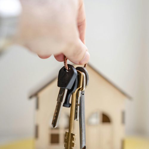 Close-up of a hand holding keys with a miniature wooden house in the background, symbolizing real estate investment.